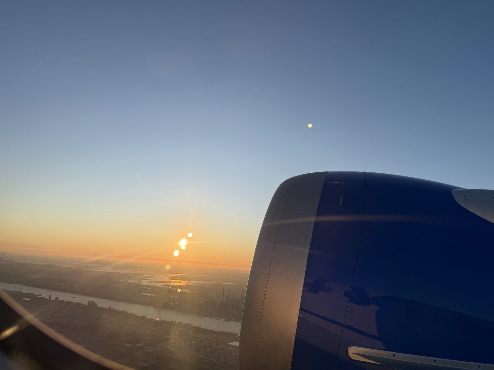 The sun rises over the New York area and the Hudson River beside a jet engine, seen from a plane window on a morning flight to California.
