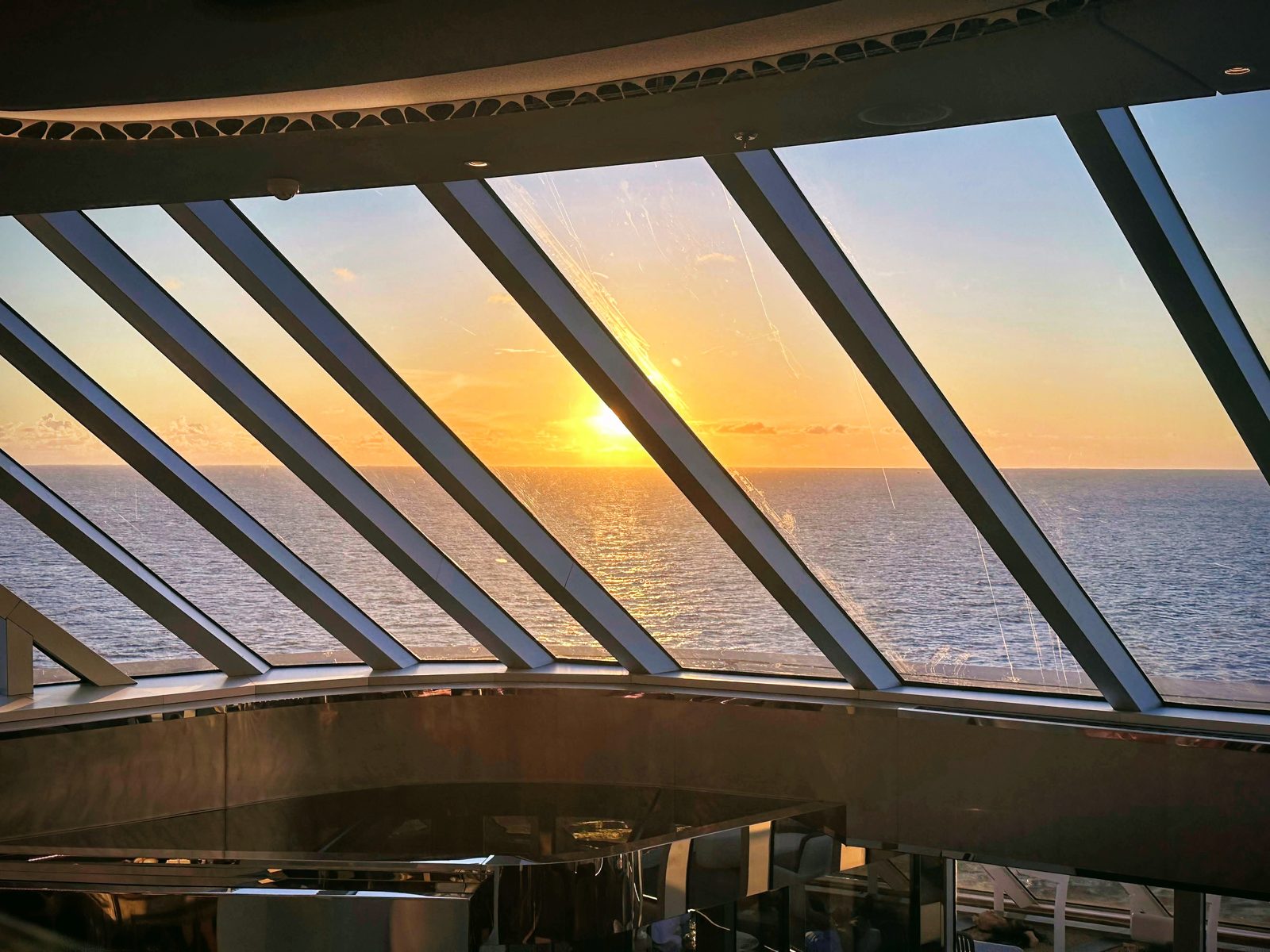 The sun sets over the ocean through the glass-panelled ceiling of a gold-toned lounge on a cruise ship.