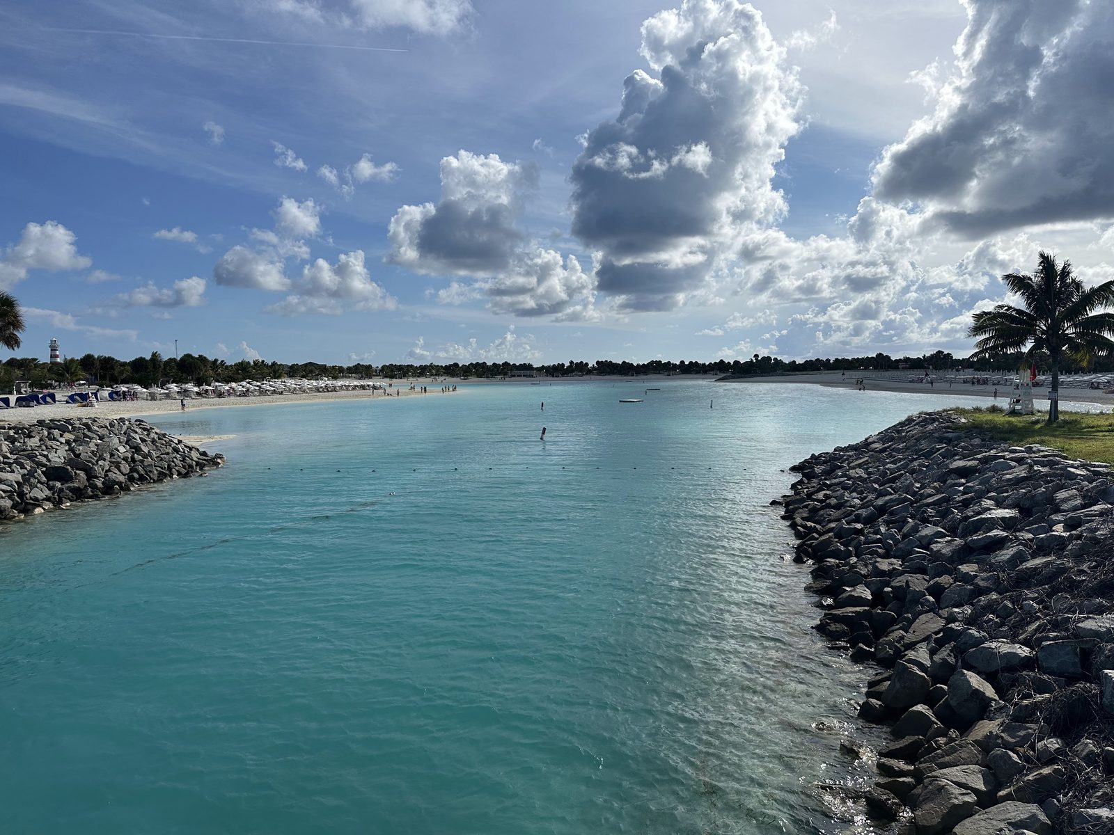 Clear turquoise water runs between a white-sand beach and a rock jetty under a bright blue sky at Ocean Cay in the Bahamas.