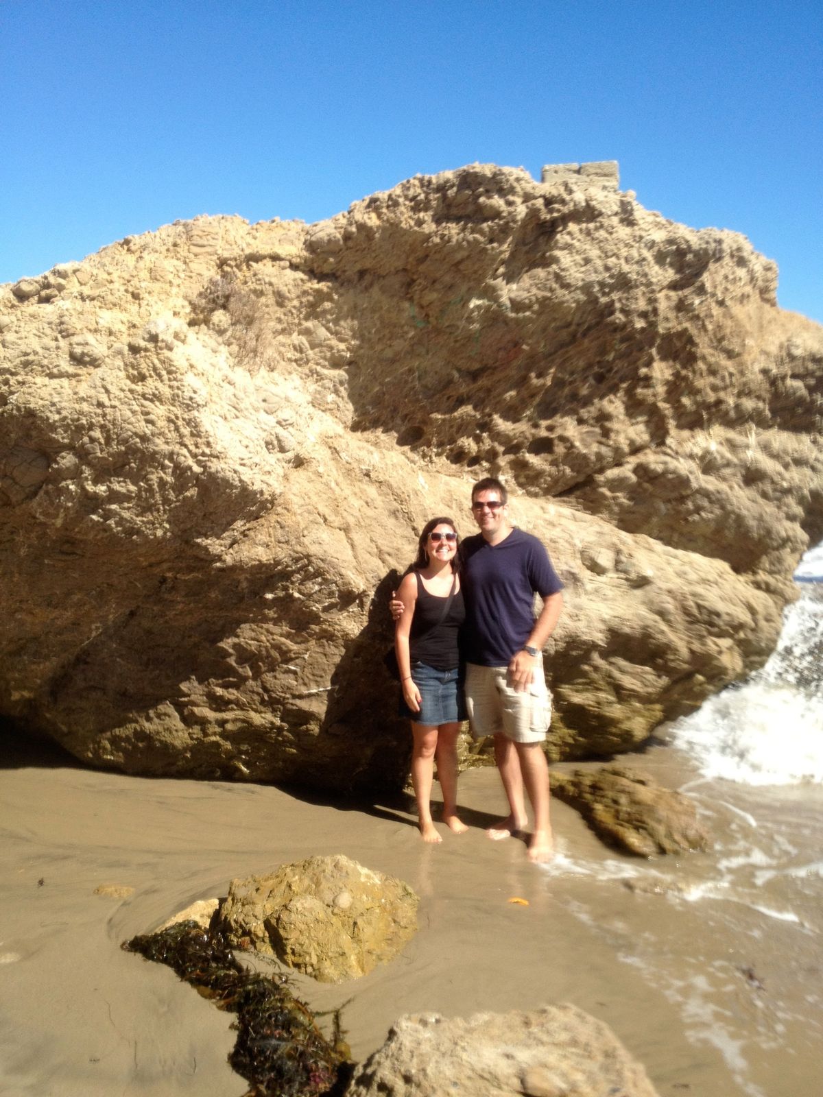 Carrolyn and Bill stand barefoot in front of a towering sandstone rock on a Southern California beach as waves roll in.