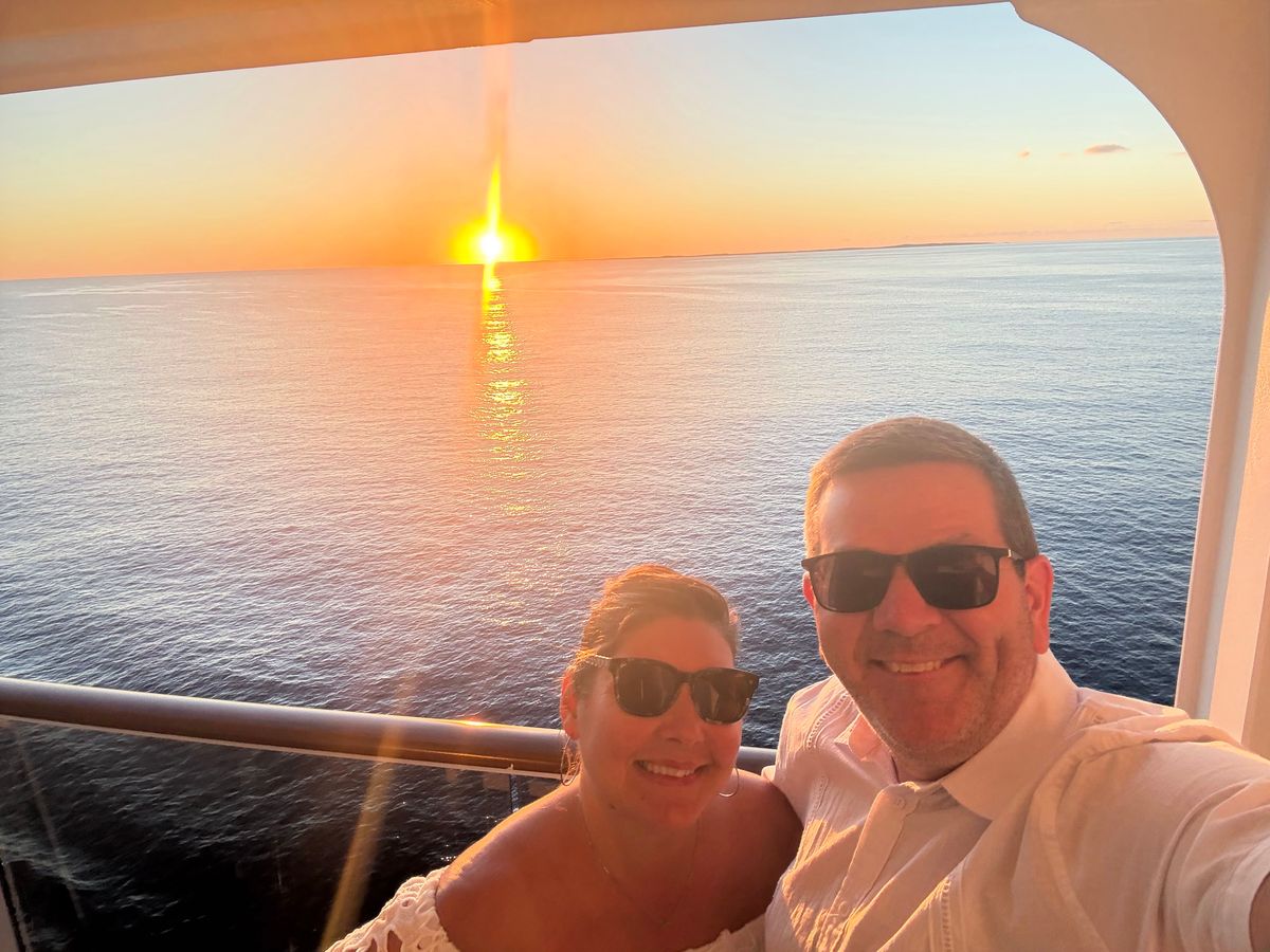 Carrolyn and Bill, in sunglasses and white summer clothes, smile for a selfie on their cruise ship balcony as the sun sets over the ocean behind them.
