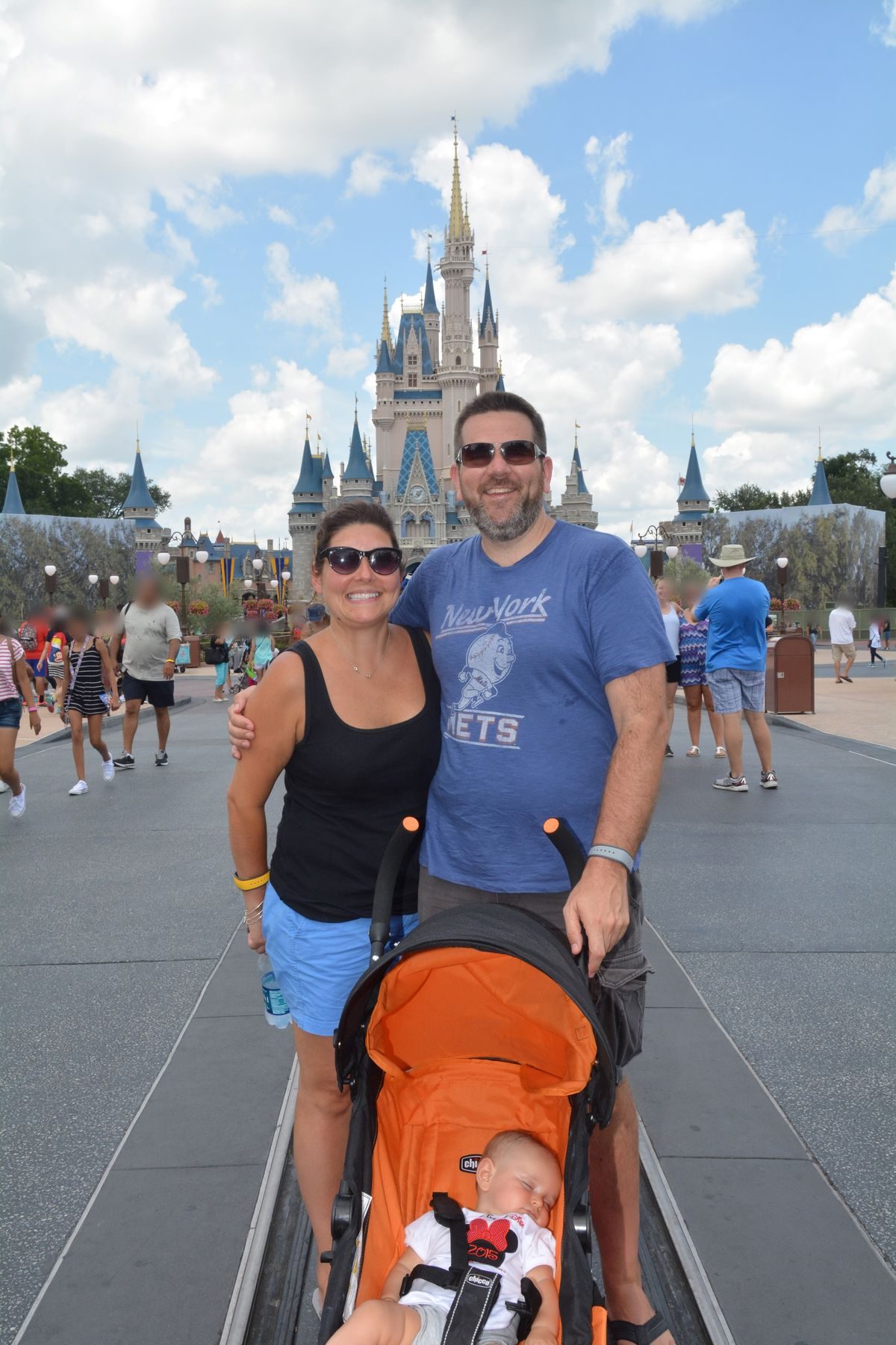 Carrolyn and Bill smile in front of Cinderella Castle at Magic Kingdom while baby Victoria naps in an orange stroller.